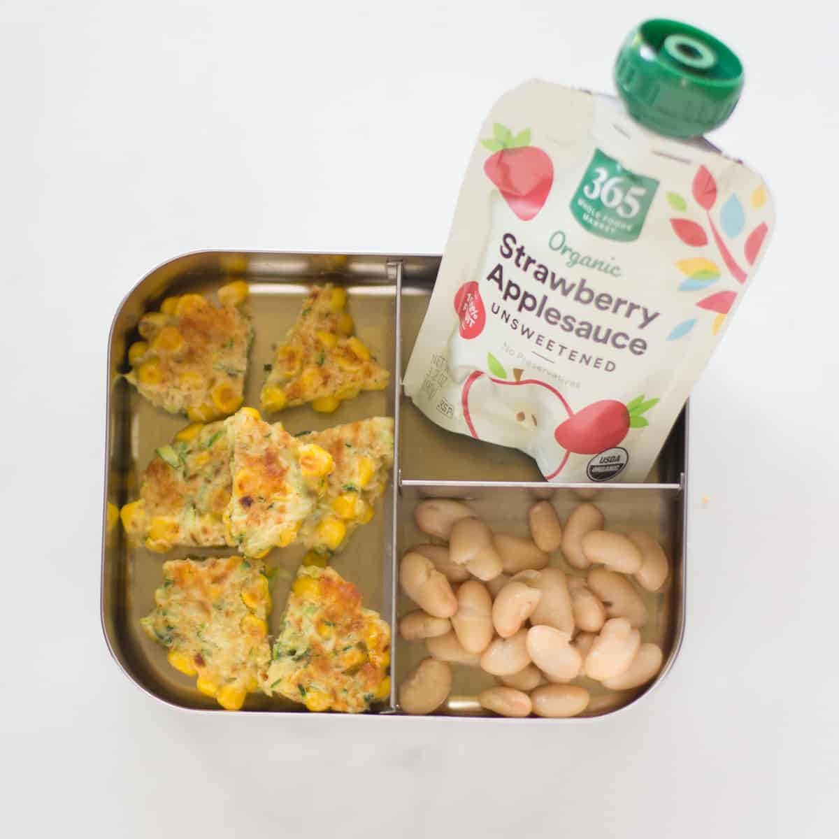 sliced fritters with white beans and applesauce pouch in a stainless steel lunch box.