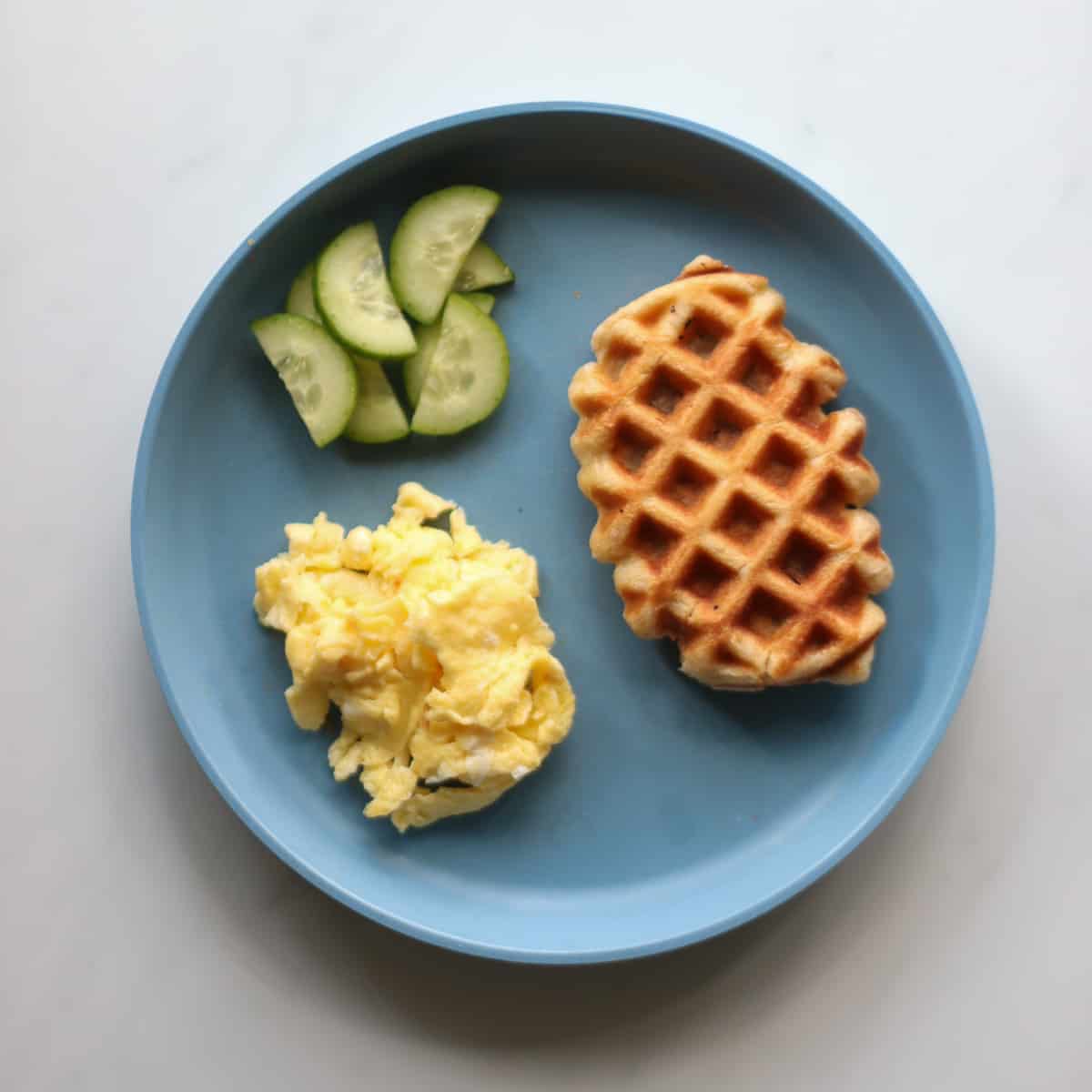 Croffle with scrambled eggs and cucumber on a blue plate.