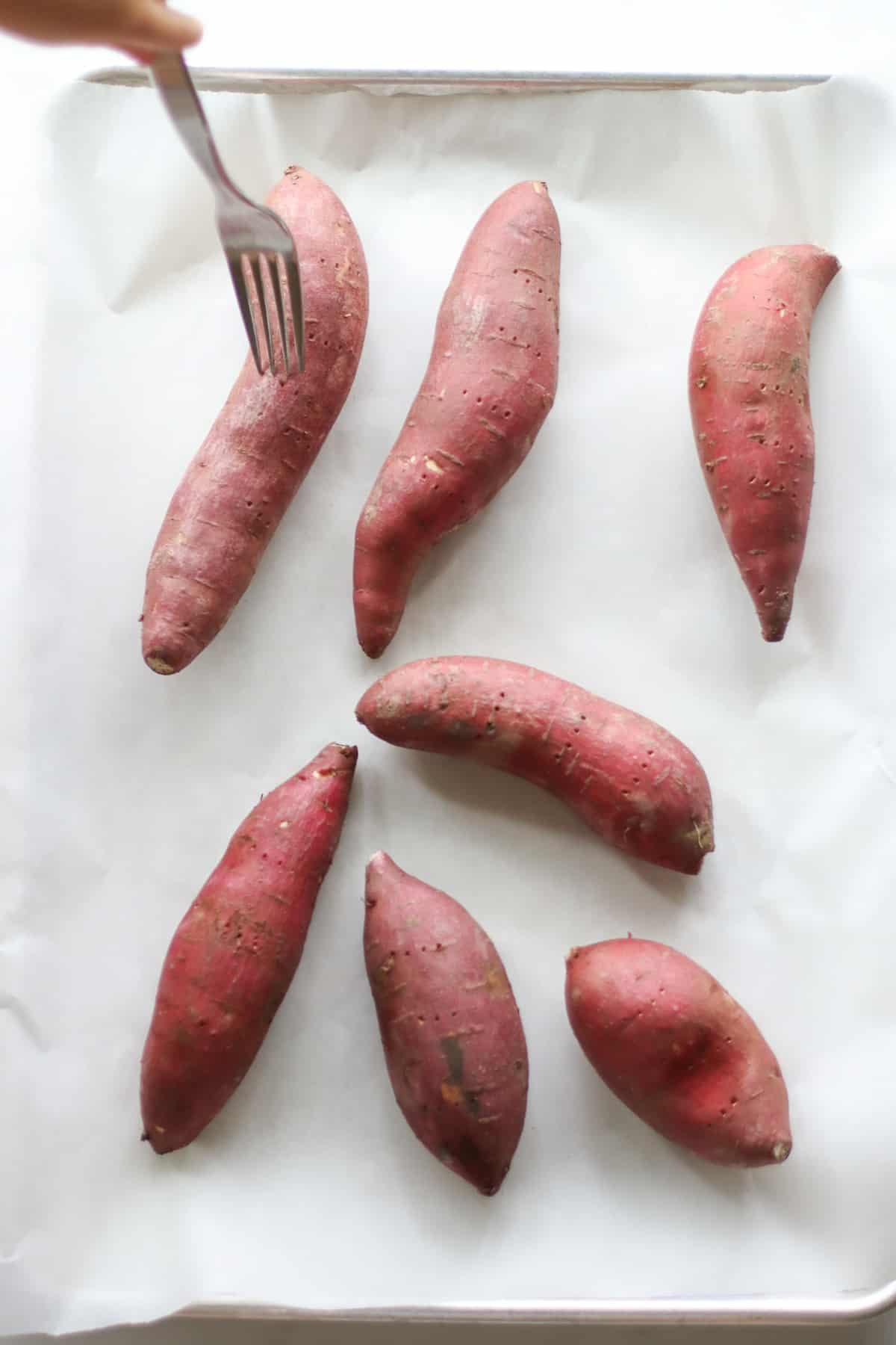 Sweet potatoes laid out on a lined baking sheet with a fork pierccing one.