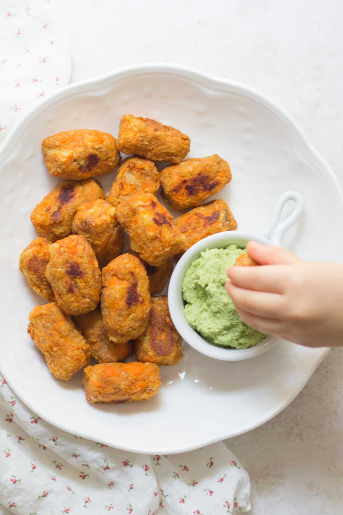 tots plated on a white plate with toddler's hand dipping one into green hummus.