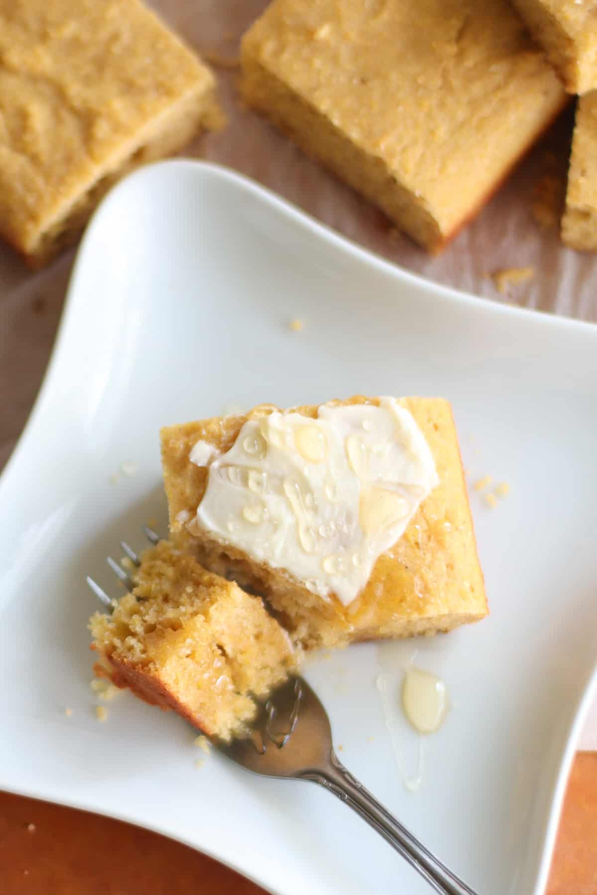 A cornbread slice with fork and topped with butter and honey.