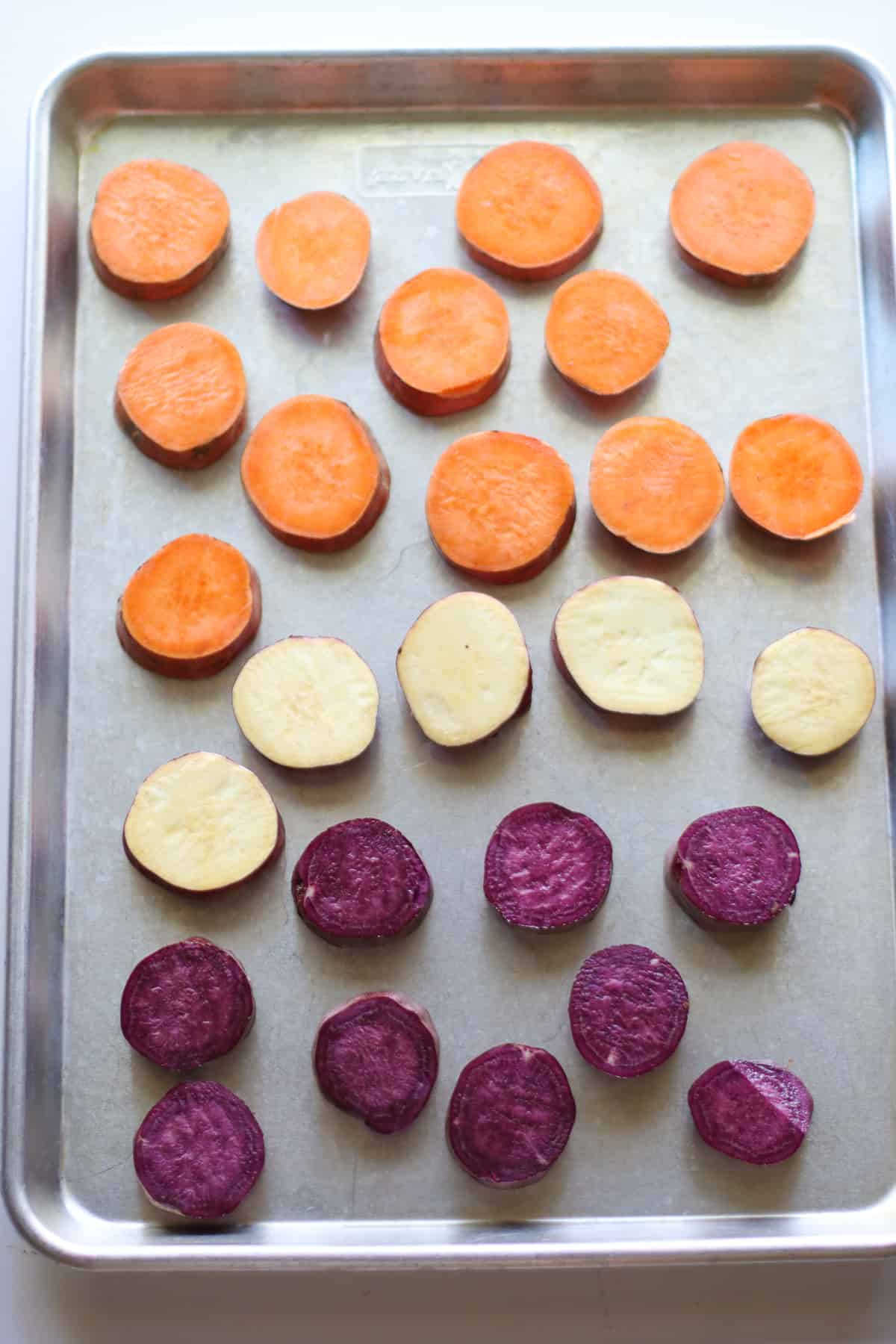 Sliced purple, orange, and white colored sweet potatoes placed on a sheet pan.
