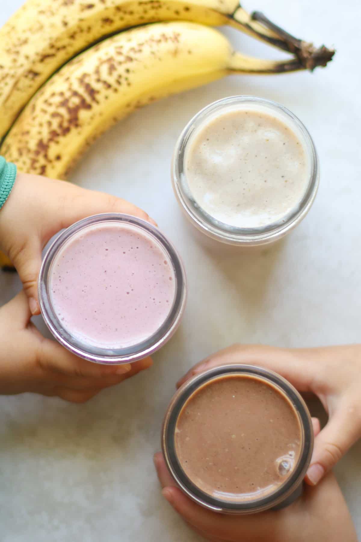 An overhead shot of 3 different flavored banana milkshakes with two toddler hands wrapped around the glass jars.