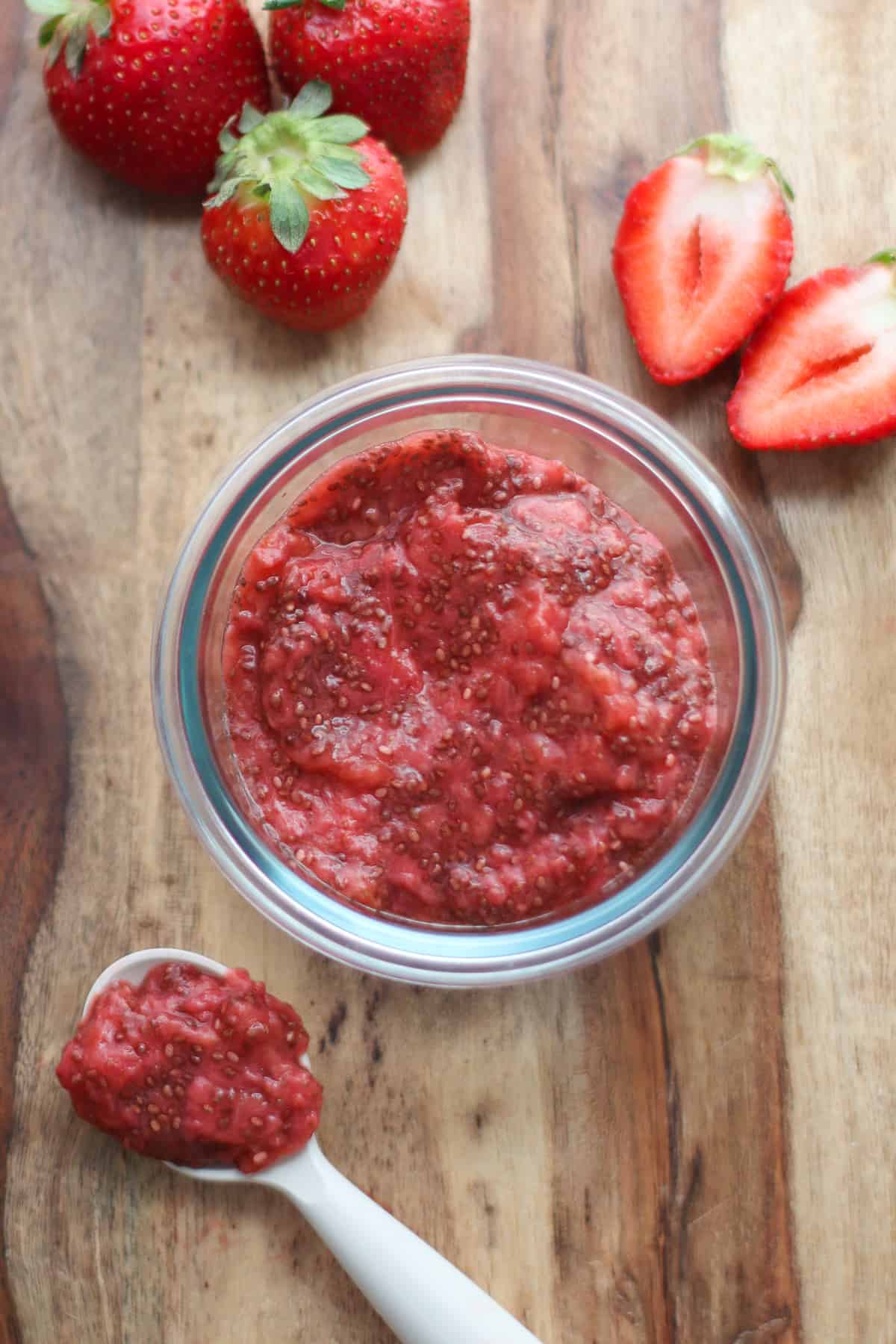 Jam stored in a glass container with fresh strawberries in the background.