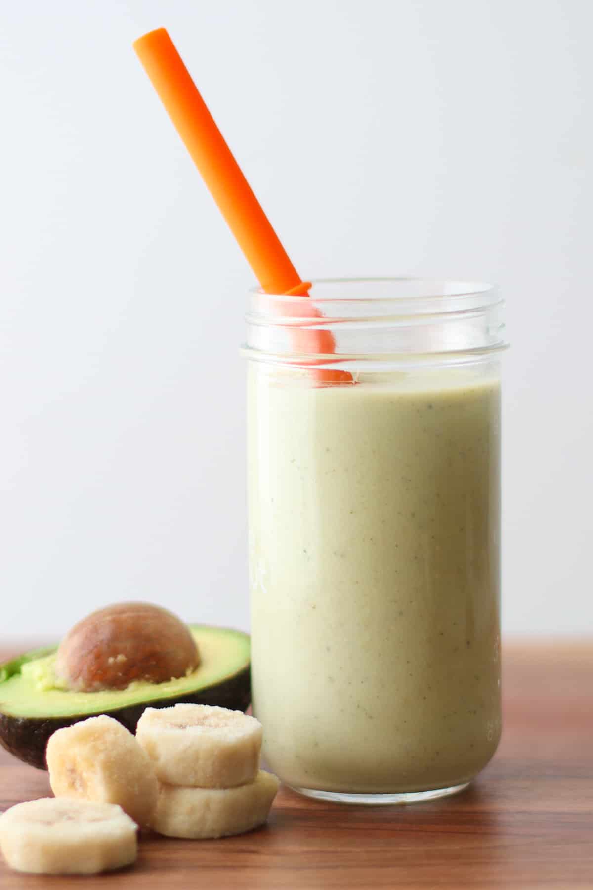 Avocado smoothie in a glass jar with a straw and bananas and avocado in the background.