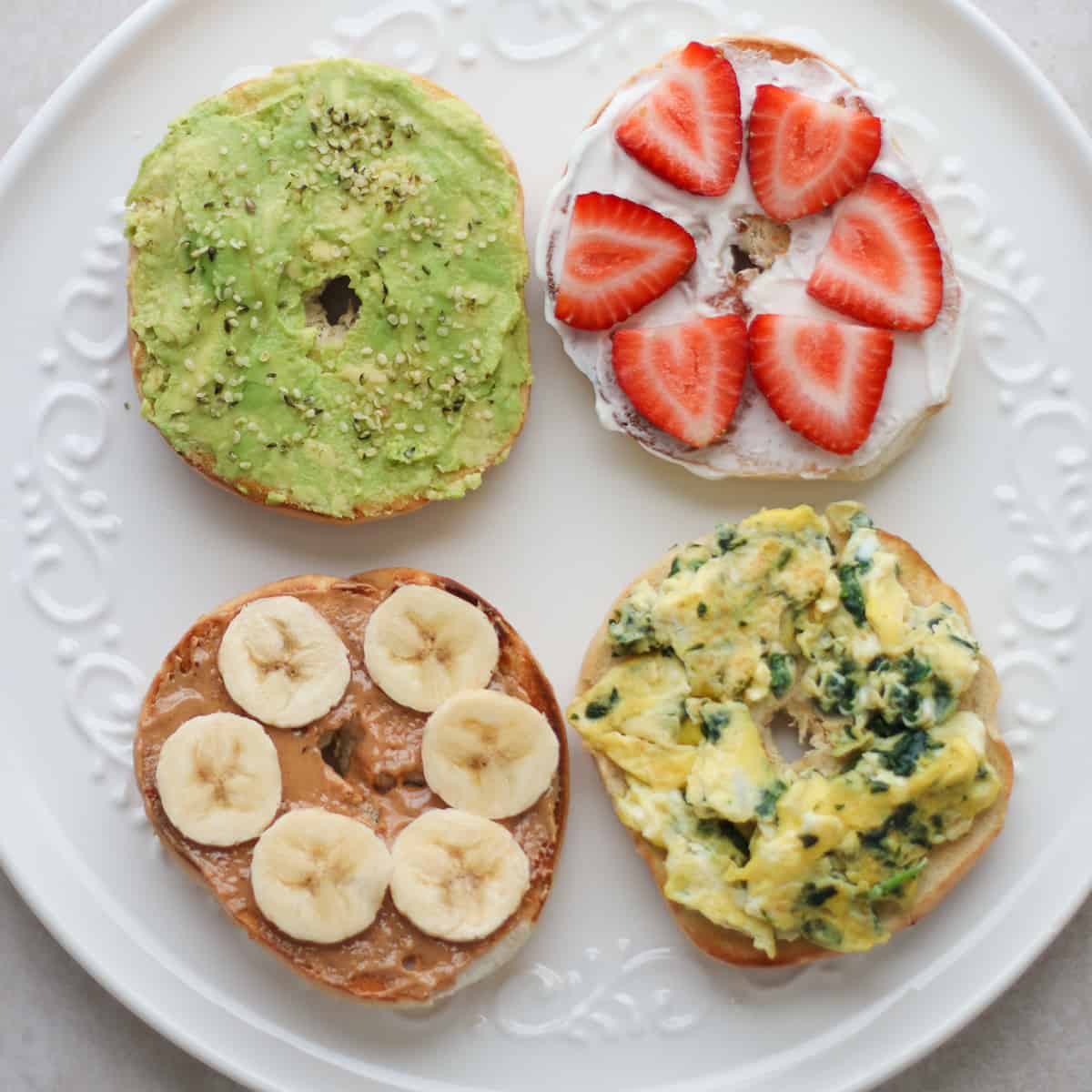 Four bagels with different toppings - avocado, peanut butter with banana, scrambled eggs, yogurt and strawberries.