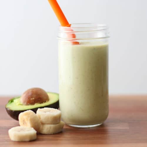 Avocado smoothie in a glass jar with a straw and bananas and avocado in the background.