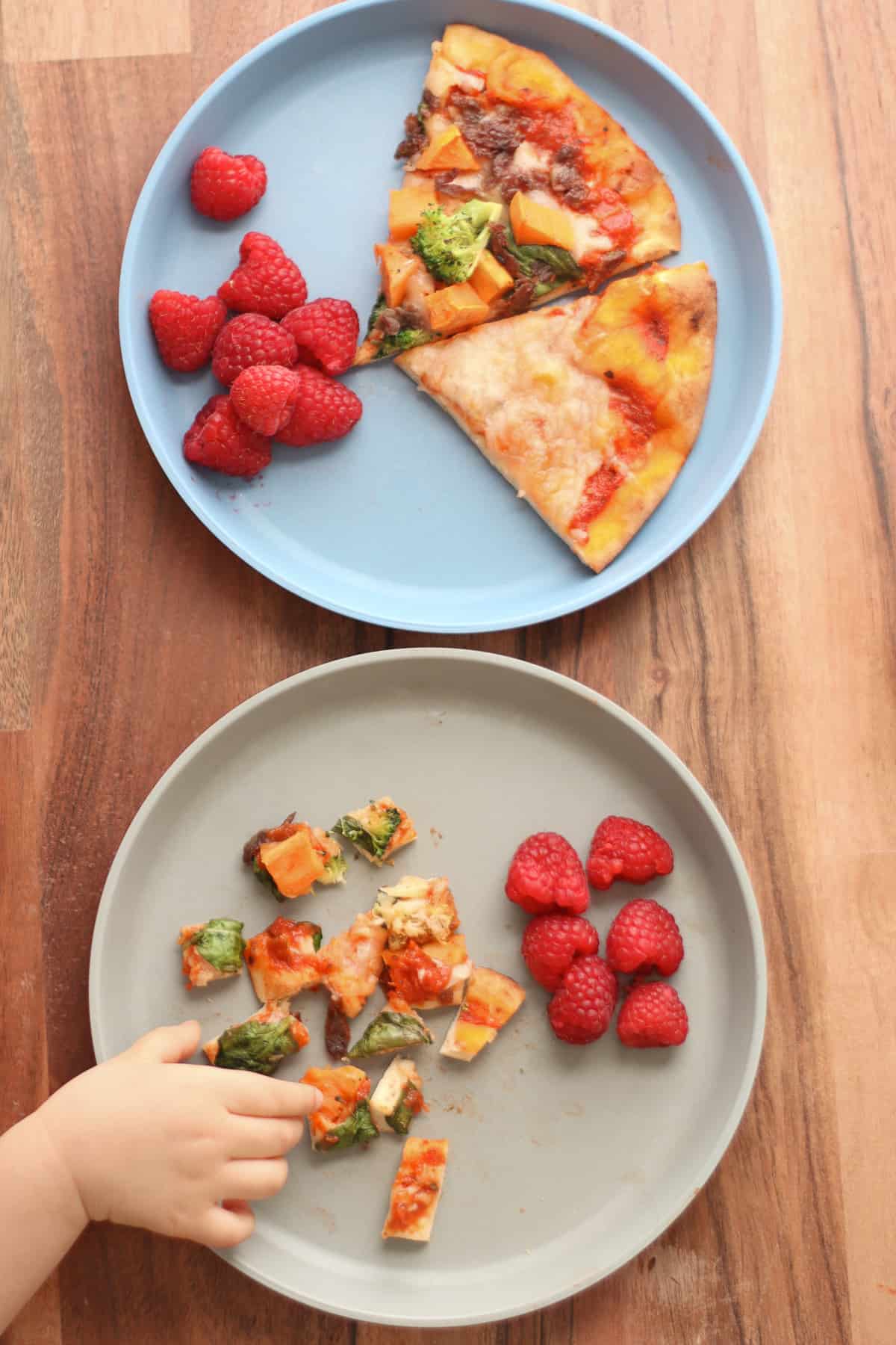 Two plates with raspberries and two pizza slices in one and bite-sized pieces in another.