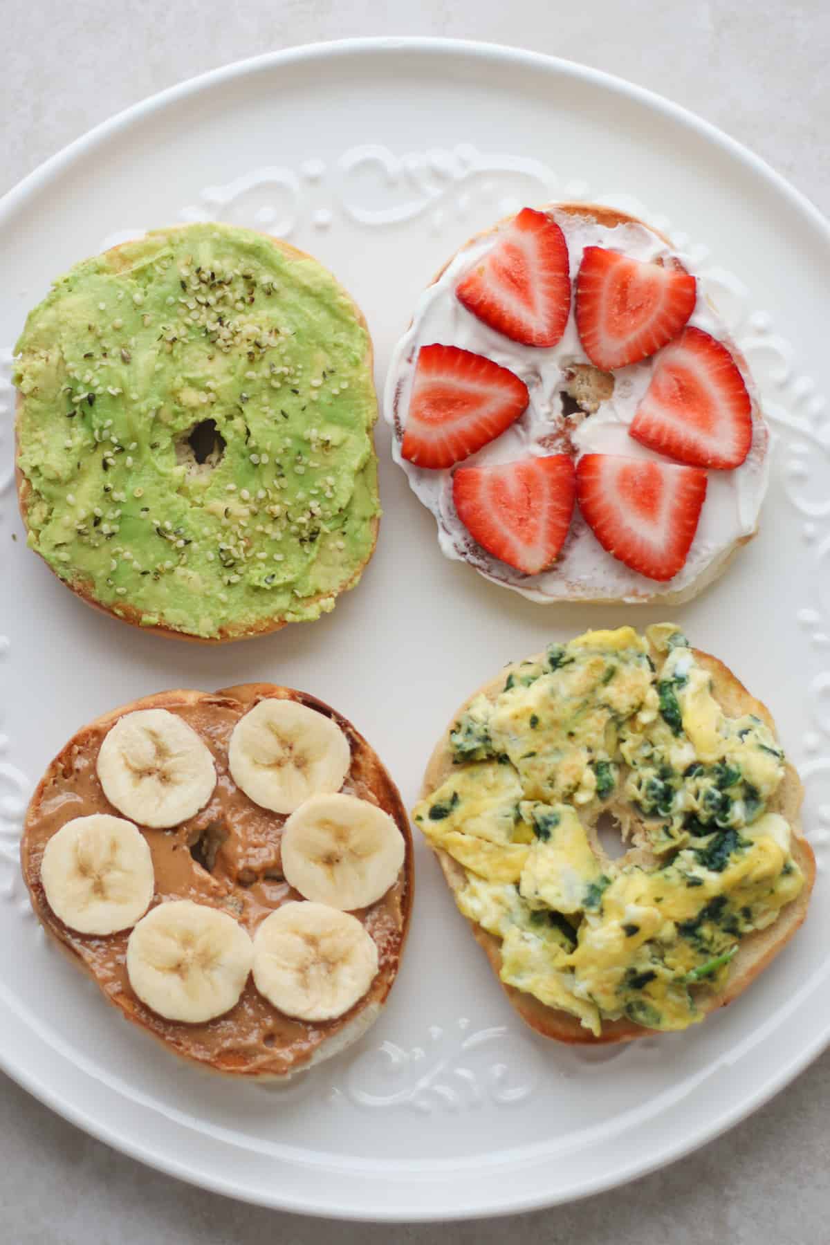 Four bagels with different toppings - avocado, peanut butter with banana, scrambled eggs, yogurt and strawberries.