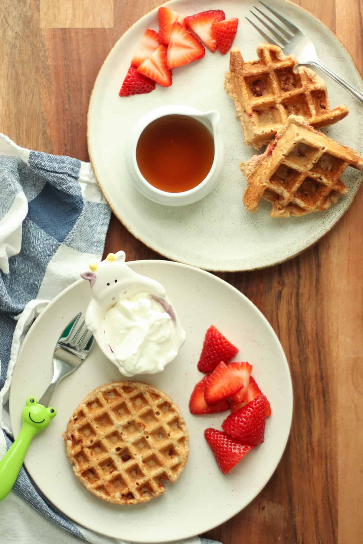Two plates with strawberry waffles, sliced berries, yogurt, and maple syrup.