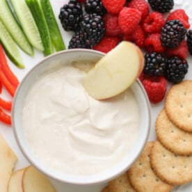 A bowl of yogurt fruit dip in the center along with fruits, veggies, crackers, and apples.