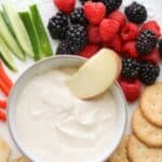 A bowl of yogurt fruit dip in the center along with fruits, veggies, crackers, and apples.
