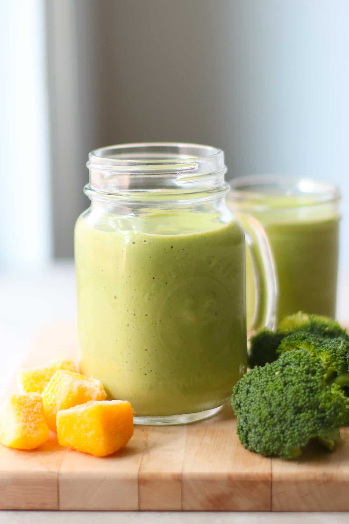 Smoothie in glass cup with mangoes and broccoli in the background.