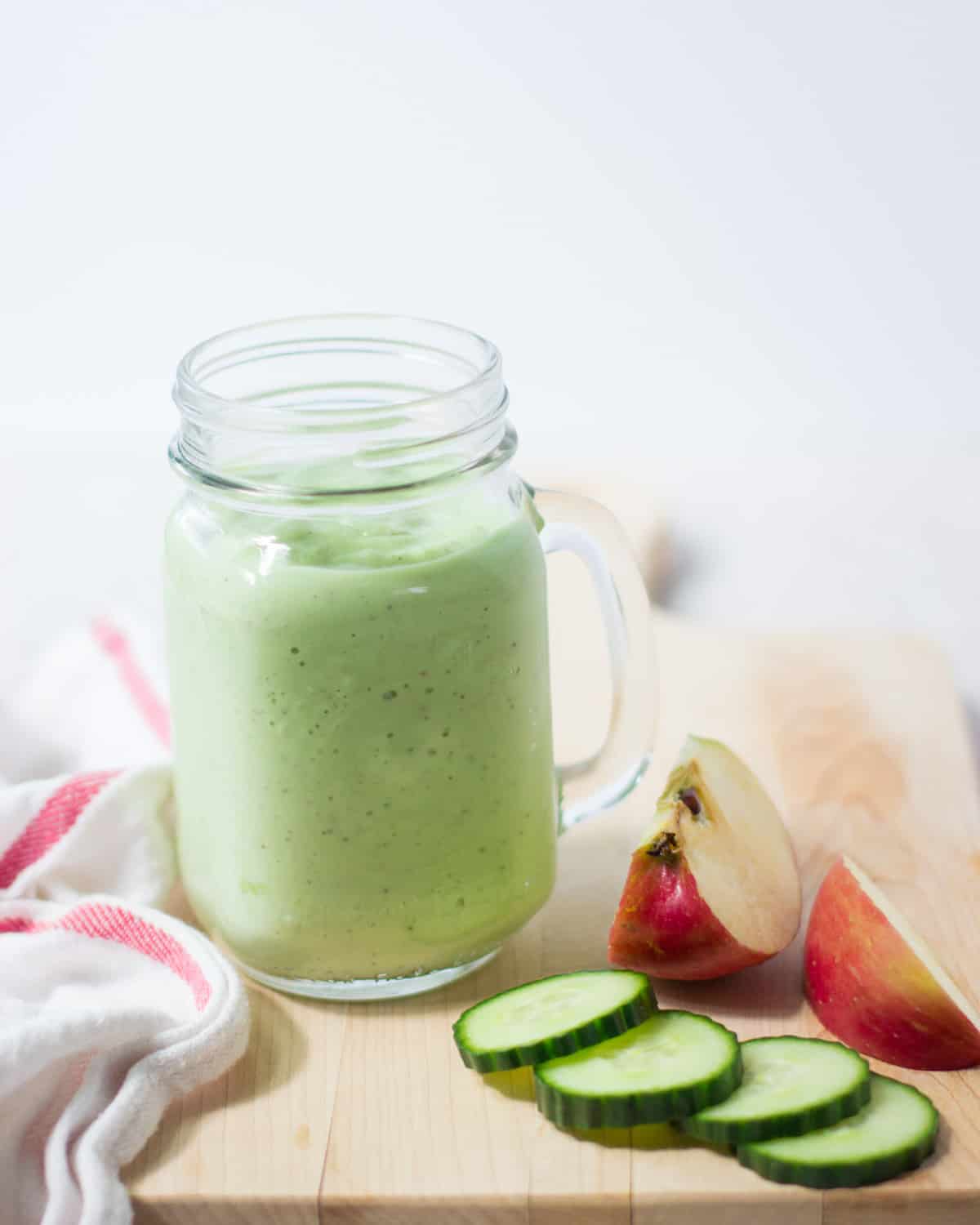 Cucumber smoothie in a glass jar with sliced cucumber and apples on the side.