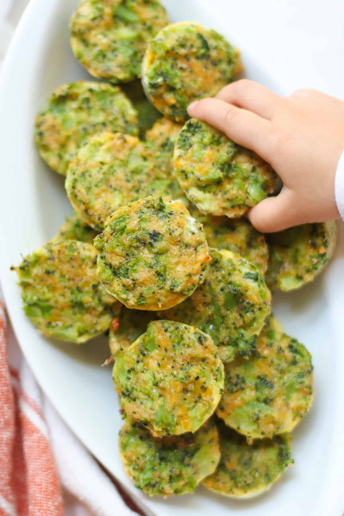 Broccoli bites piled on high on a white plate with a toddler's hand.