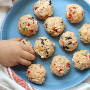 An overhead shot of chicken rice balls on a blue plate with toddler's hand grabbing one.
