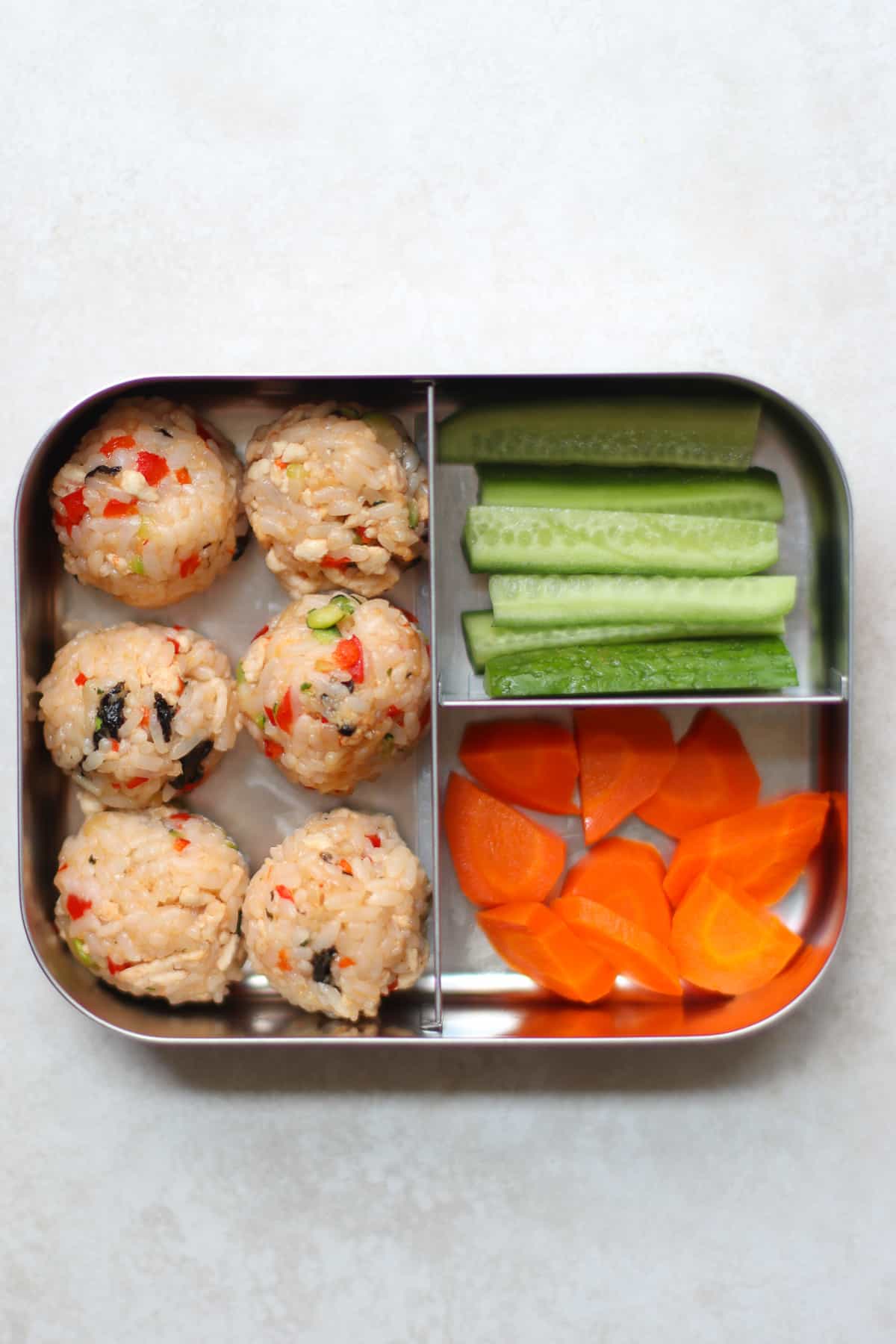 Six rice balls in a stainless steel lunchbox with cucumbers and carrots.
