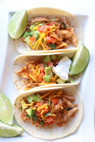 3 chicken tacos in a white plate with lime wedges.