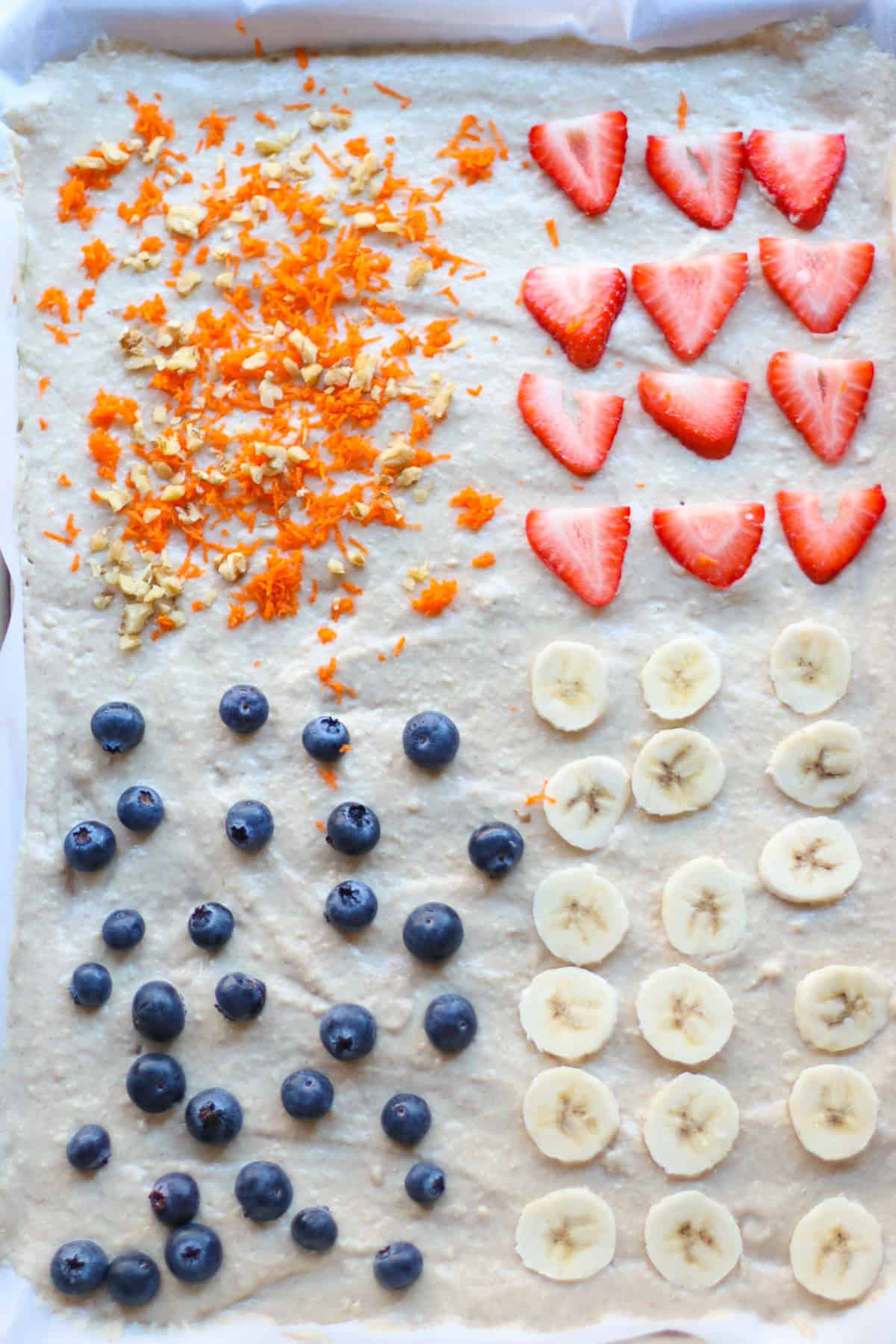 Unbaked sheet pan pancakes in a sheet pan, one-fourth with grated carrots and chopped nuts, one-fourth with sliced strawberries, one-fourth with whole bluerries and one-fourth with sliced bananas.