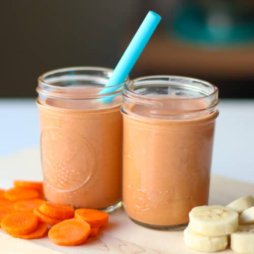 Two mason jars, one with a blue straw, filled with the smoothie on a wooden cutting board with sliced carrots and sliced banana.