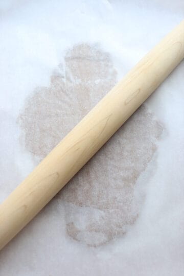 Dough rolled between parchment paper with a wooden rolling pin on top.