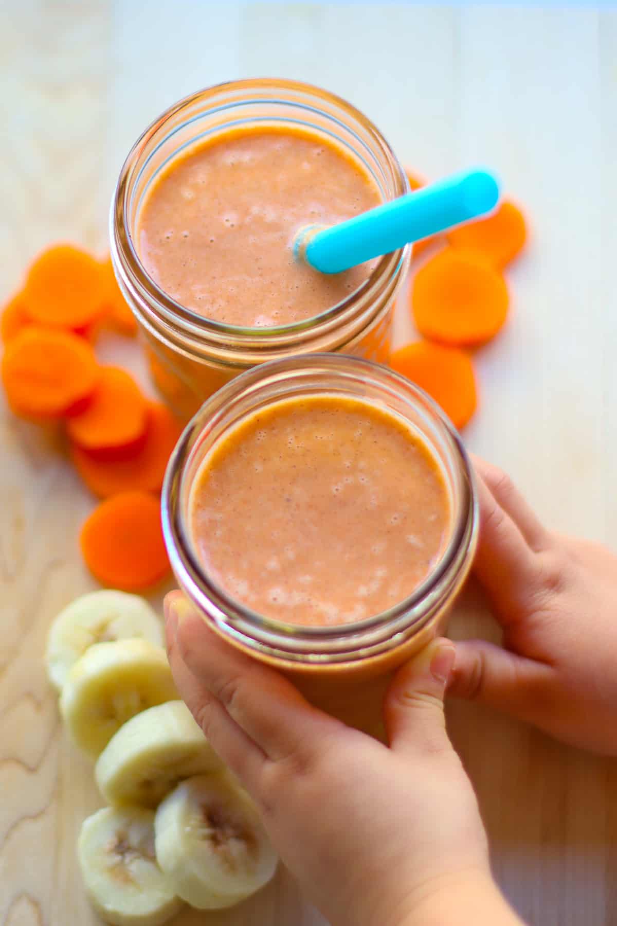 Top view of two smoothies in a mason jar, one with a blue straw, surrounded by slices of carrots and bananas.