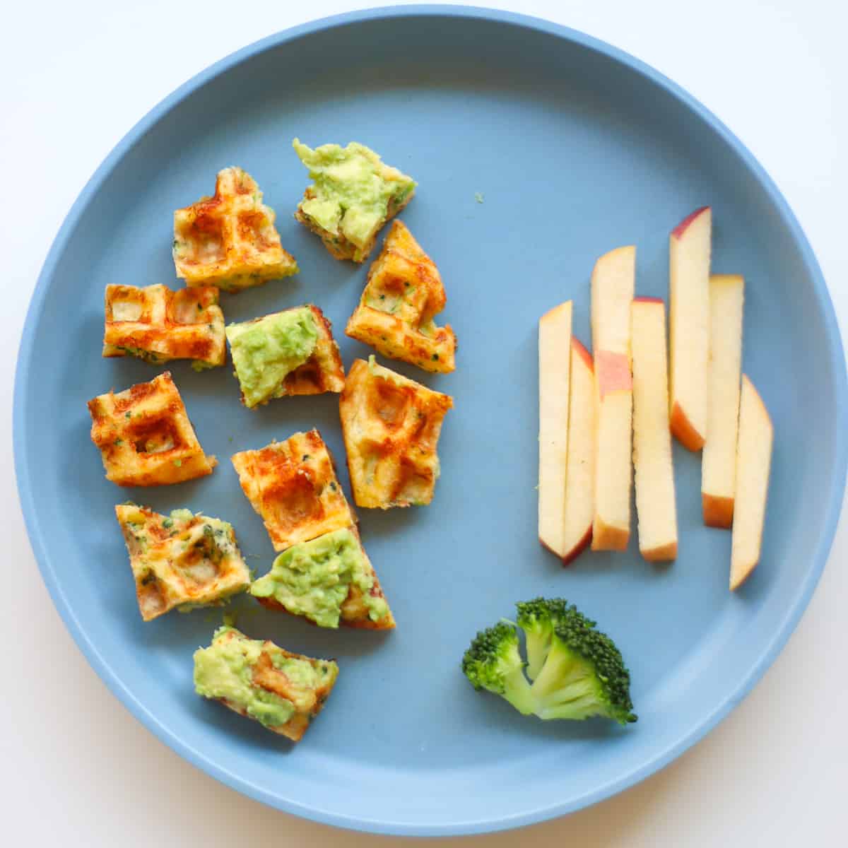 A blue plate with a waffle cut into pieces served with a piece of broccoli and apple spears.