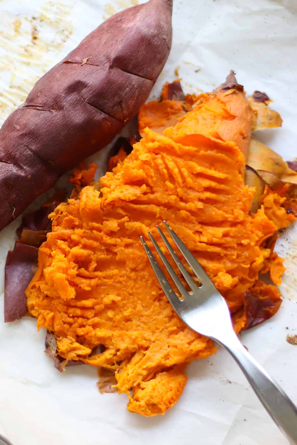 A sweet potato being mashed with a fork on parchment paper.