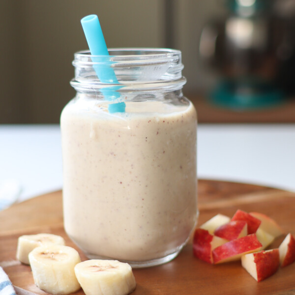 Blended apple banana smoothie in a mason jar with a blue straw on a wooden cutting board surrounded by slices of banana and apple.