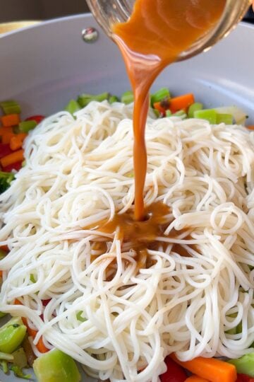 Sauce being poured over noodles and vegetables in a skillet.