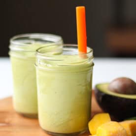 Two clear mason jars with finished mango avocado smoothies, one with an orange straw in it, along with mango chunks and half of an avocado.