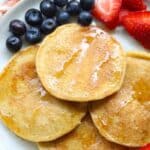 Four cooked pancakes layered on a white plate drizzled with syrup served along with fresh blueberries and sliced fresh strawberries.