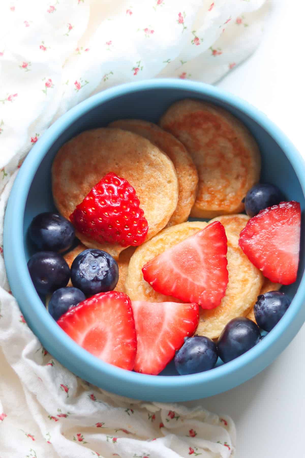 Pancakes in a blue serving dish topped with fresh sliced strawberries and fresh blueberries.