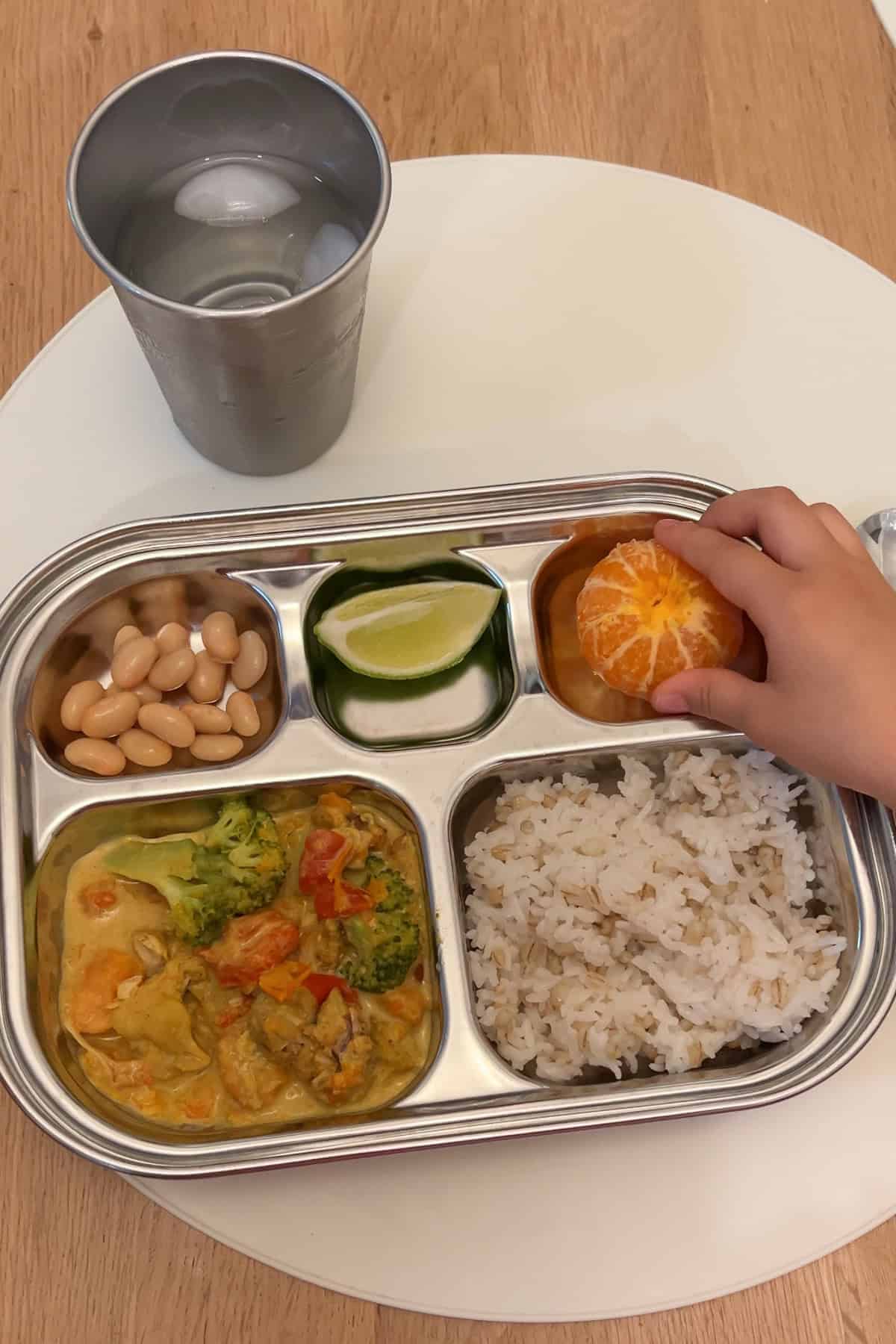 A kids 5-compartment silver metal plate with a serving of the chicken curry in the bottom left compartment, a serving of rice in the bottom right compartment, white beans in the top left, a lime wedge in the top middle, and a peeled mandarin orange in the tip right, served with a glass of water in a metal cup.