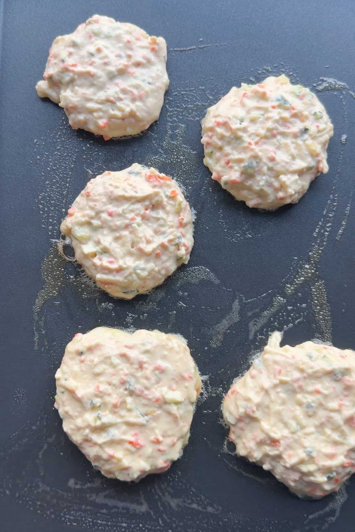 A lightly greased skillet with 5 scoops of pancake batter spread slightly.