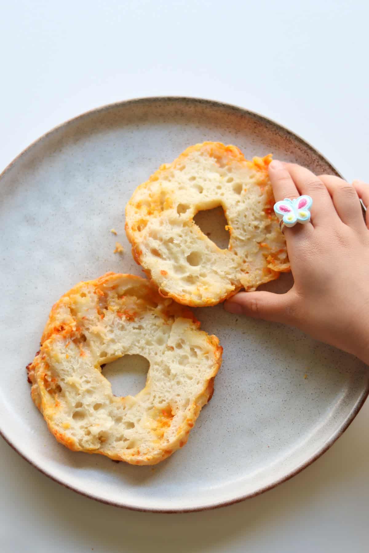 Sliced bagel on a plate with a toddler hand grabbing it.