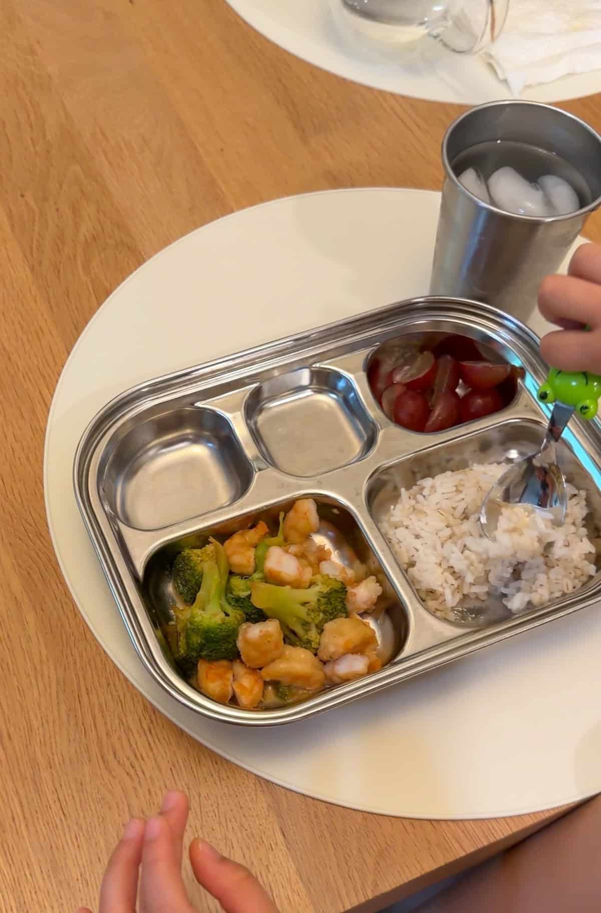 Toddler's stainless steel plate with shrimp broccoli stir fry, rice, and grapes.