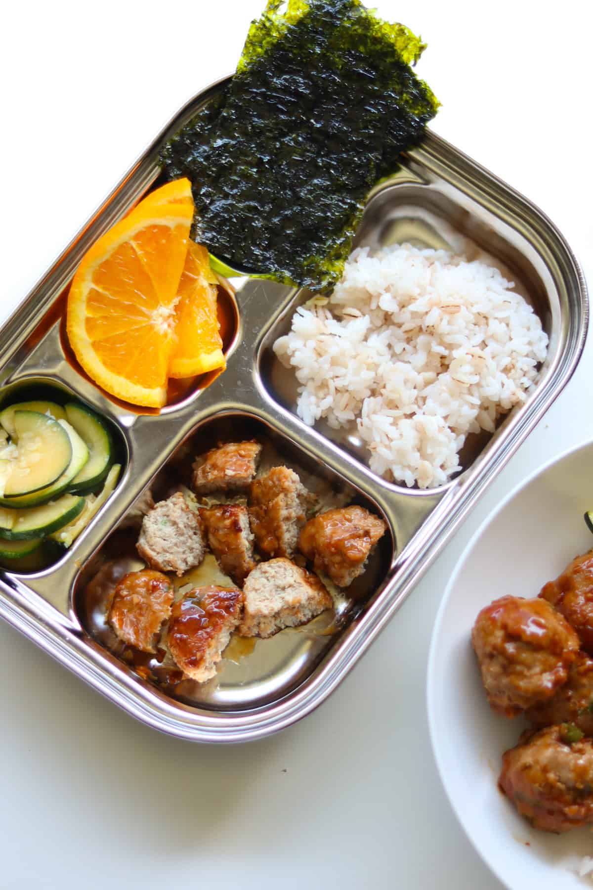 Teriyaki meatballs cut into small pieces with rice, orange, seaweed snack, and cucumber in a stainless steel plate.