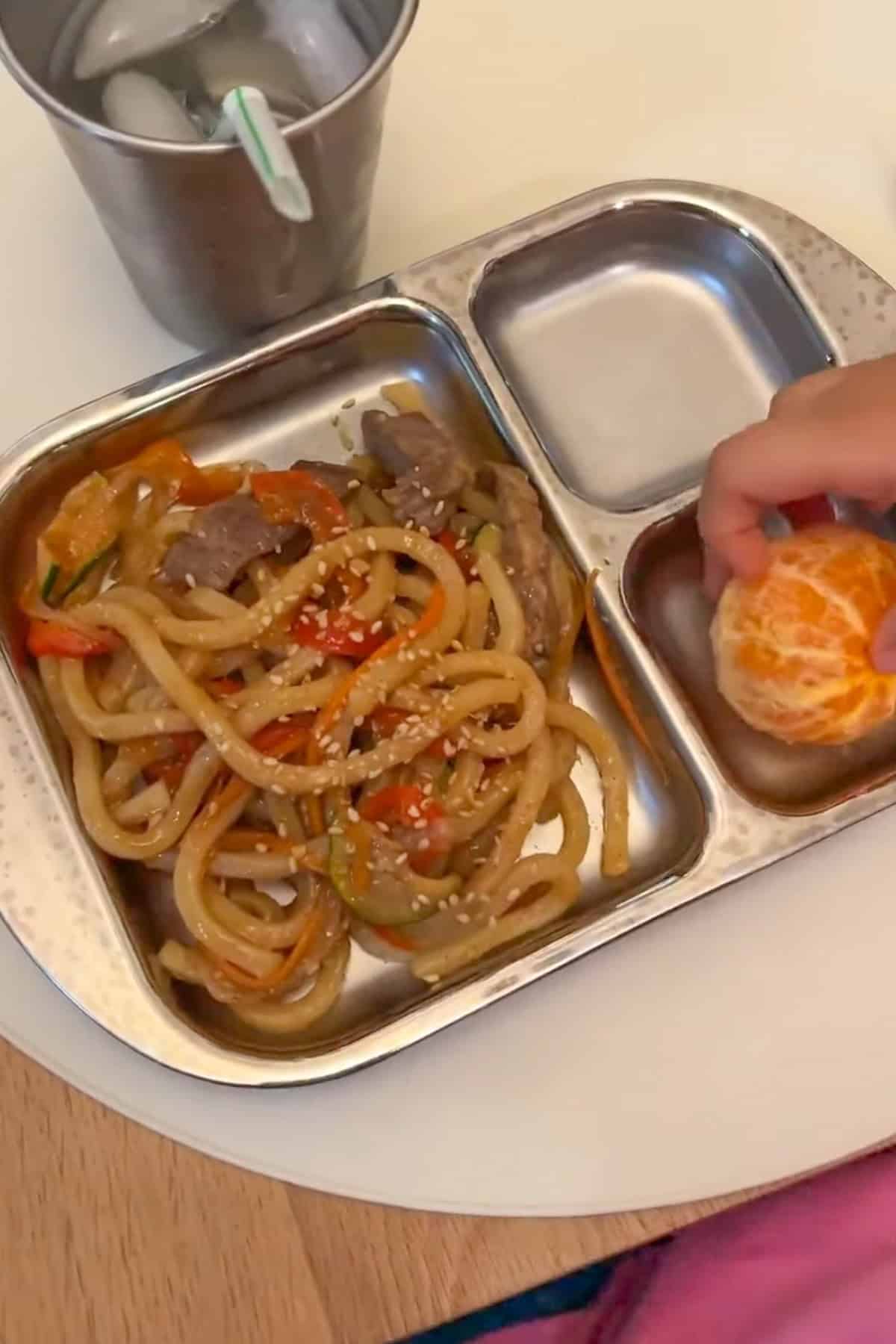 beef udon stir fry served on a divided stainless steel plate with orange.